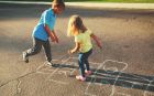 Zwei Kinder spielen bei Sonnenschein auf einer Strasse ein Hüpfspiel, das mit Kreide auf den Asphalt gezeichnet wurde.